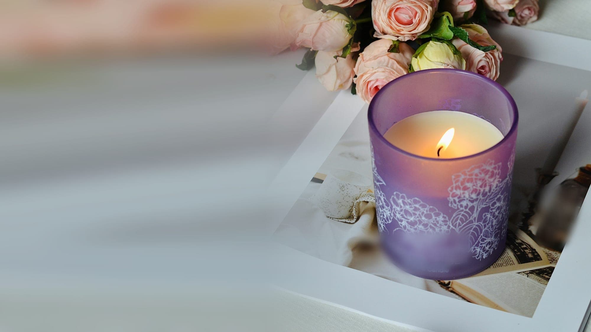 A lit candle in a lilac glass jar beneath a spray of blush roses