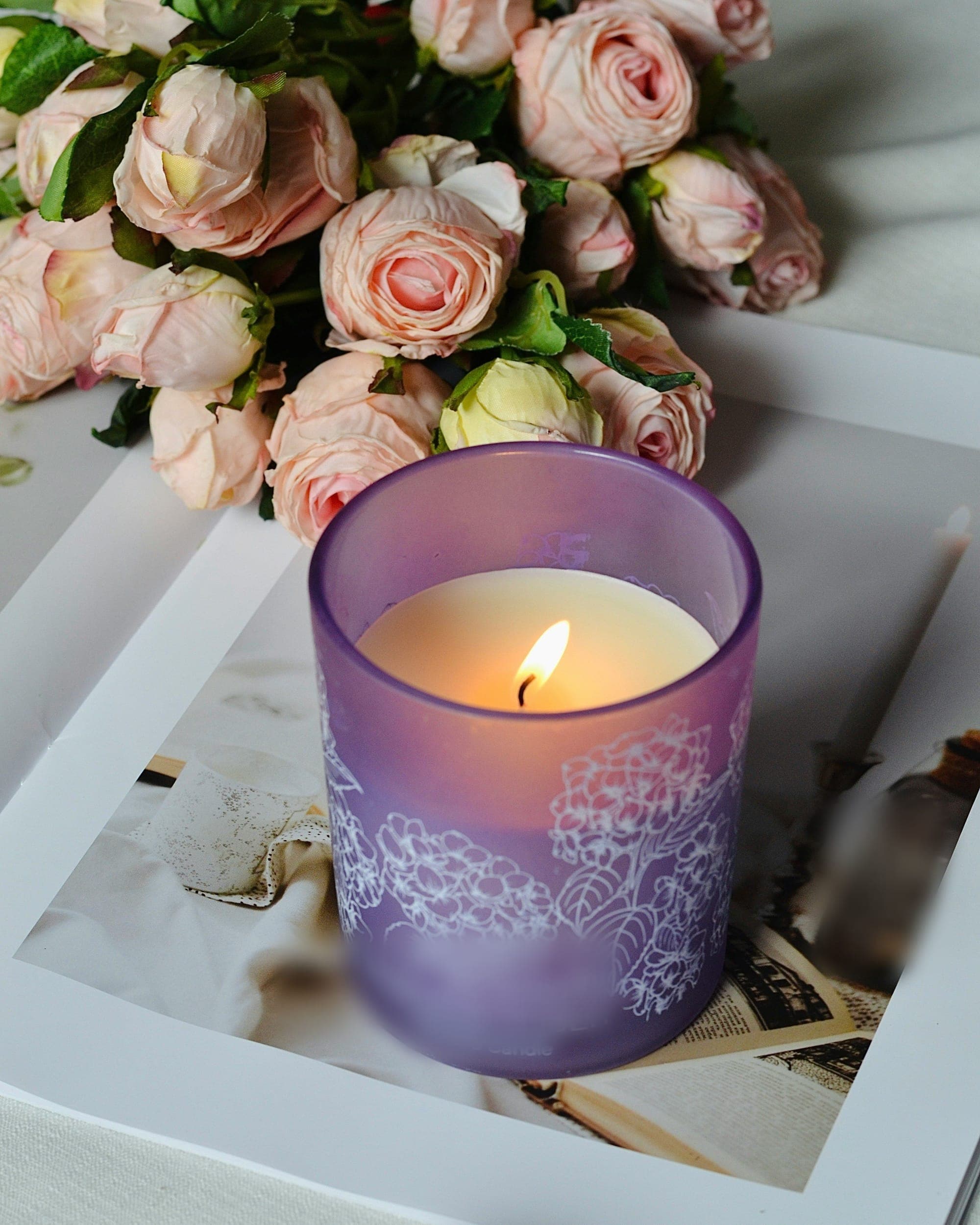 A lit candle in a lilac glass jar beneath a spray of blush roses
